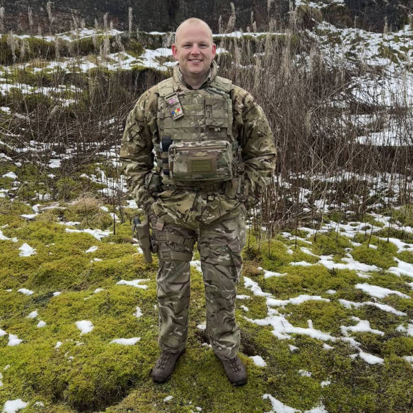 A soldier in camouflage gear stands on mossy ground with patches of snow, holding equipment.