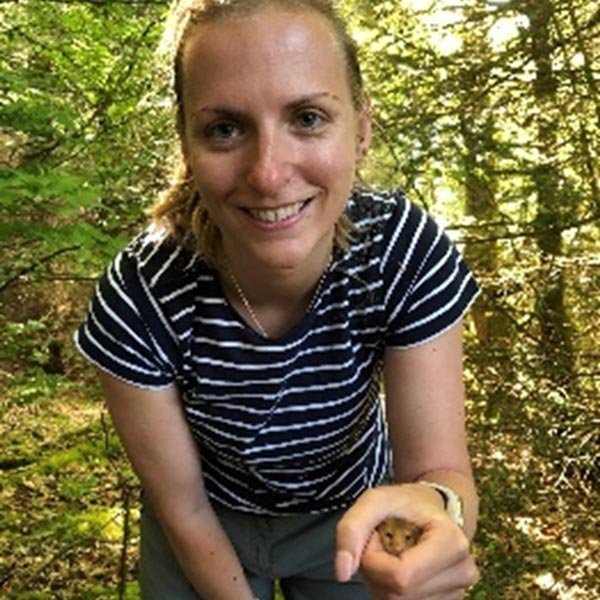 A person in a striped shirt poses outdoors in a forest, with greenery surrounding them holding a small mouse.