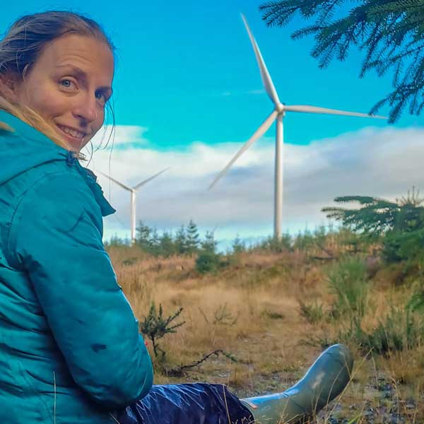 A person in a blue jacket sits on the ground, surrounded by wind turbines and green trees under a bright blue sky.