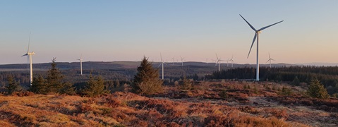 A landscape featuring multiple wind turbines across rolling hills, surrounded by patches of grass and trees under a clear sky.