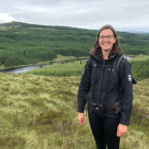 A person stands outdoors in a black jacket, surrounded by green hills, trees, and a winding river under a cloudy sky.