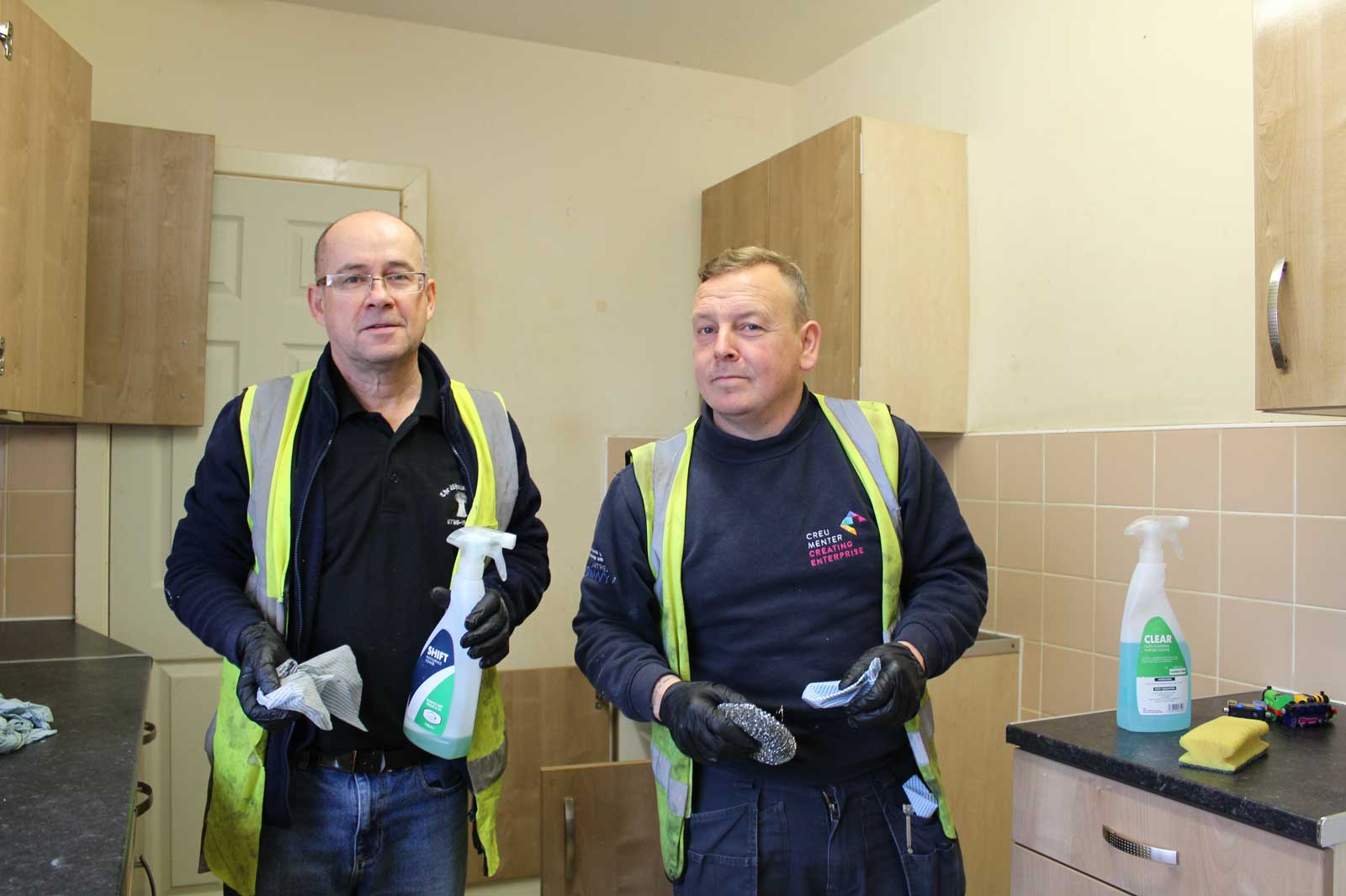 Two workers in a kitchen holding cleaning supplies, wearing safety vests, with cabinets in the background.