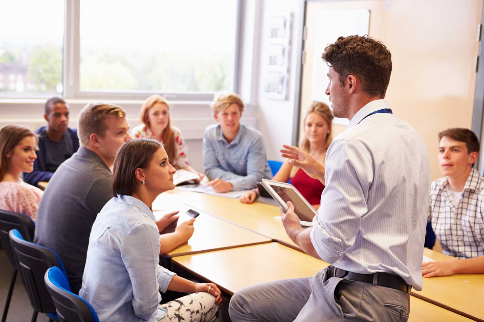 A group discussion in a classroom setting, featuring a presenter engaging with attendees seated around a table.