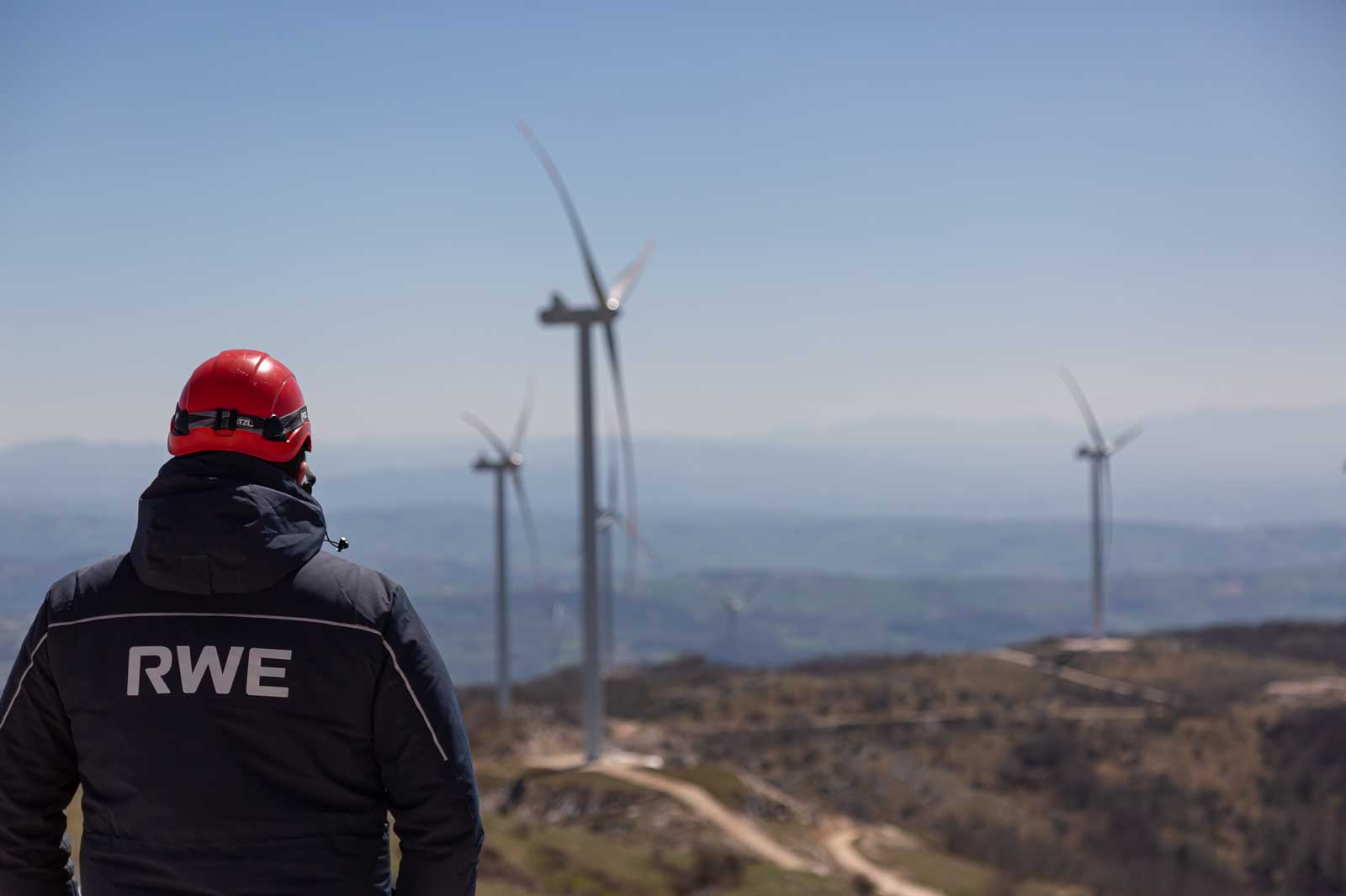 A technician in a red helmet gazes at wind turbines on a sunny day, showcasing renewable energy in a mountainous landscape.