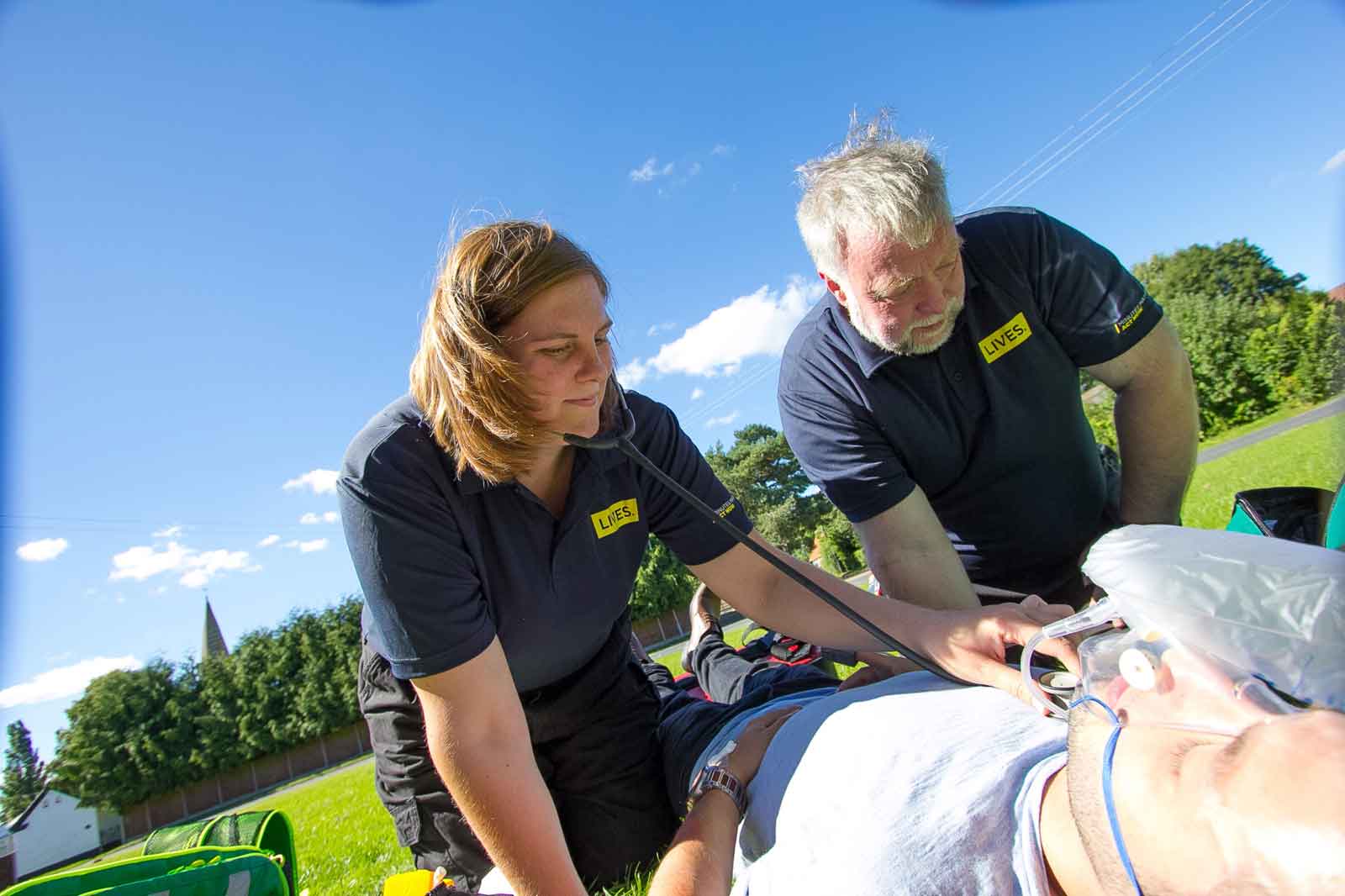 A man and woman in dark t shirts are kneeled infront of a man lying on the floor with a oxygen mask on taking his heartbeat