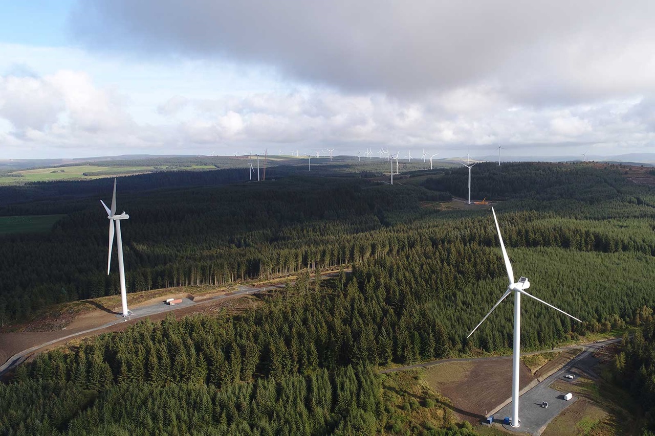 Aerial view of wind turbines amidst lush green forests under a cloudy sky.