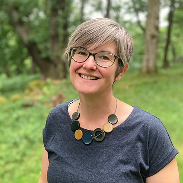 A woman with short grey hair wears a dark t-shirt and a colourful necklace in a green outdoor setting.