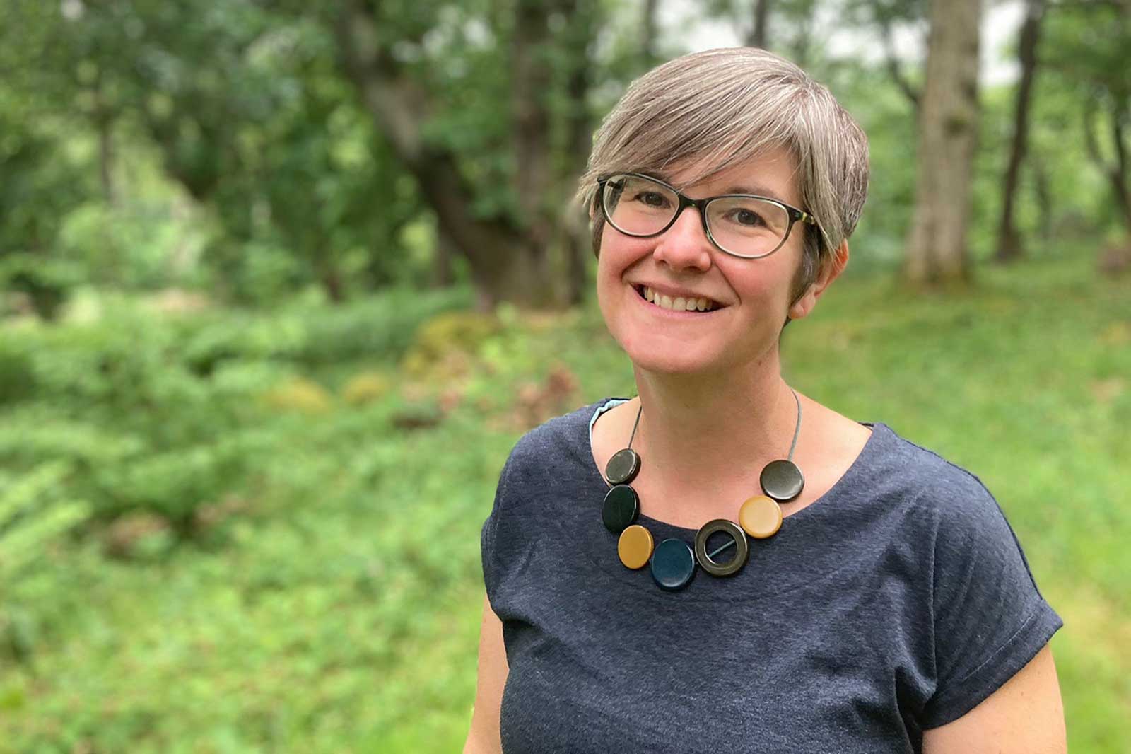 A woman in a dark shirt, wearing a distinctive necklace, stands in a lush green forest setting. Portrait of Katy Woodington.