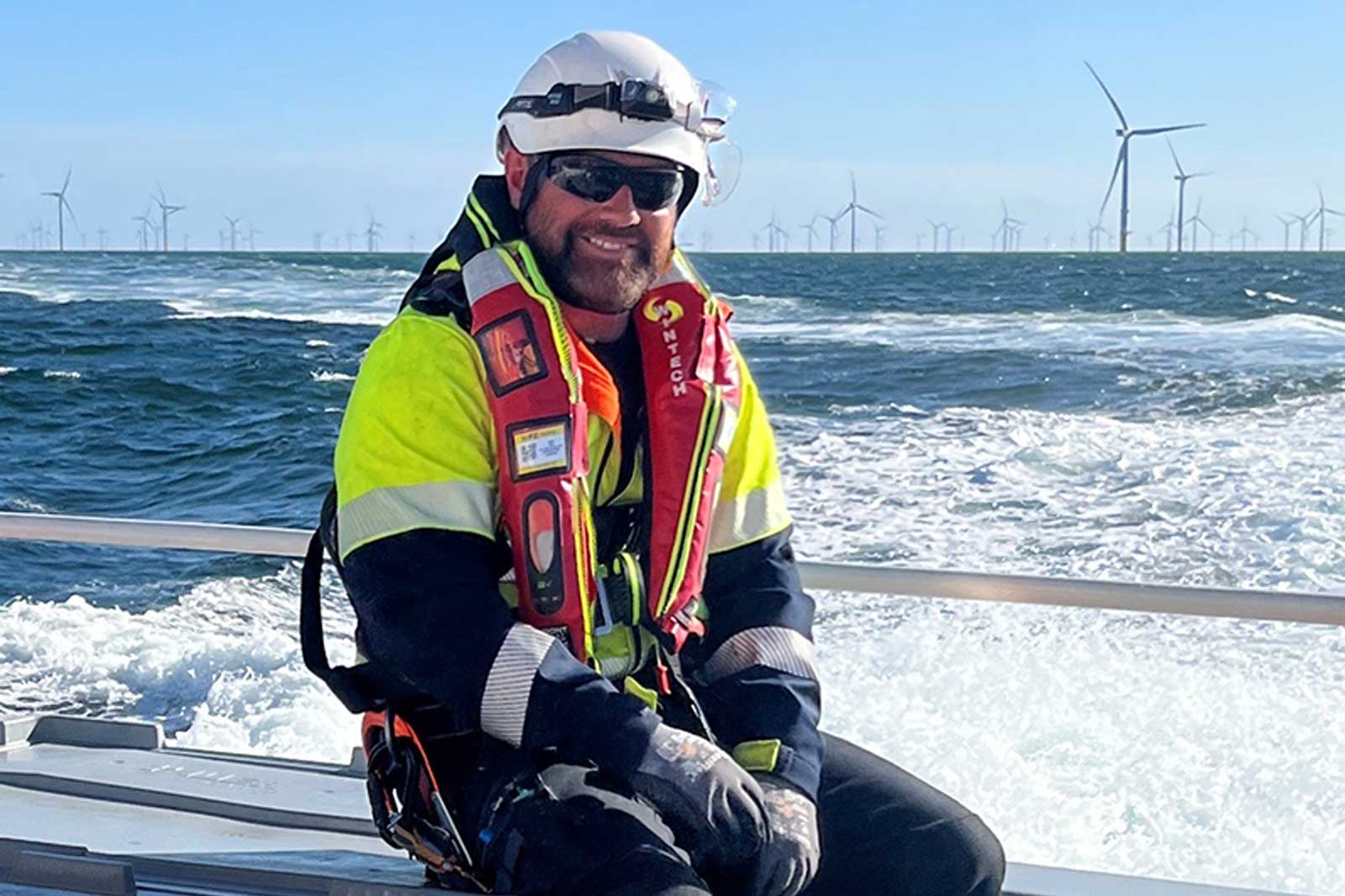 A worker in a high-visibility jacket and life vest sits on a boat with wind turbines in the background. Portrait of Stewart Harris.
