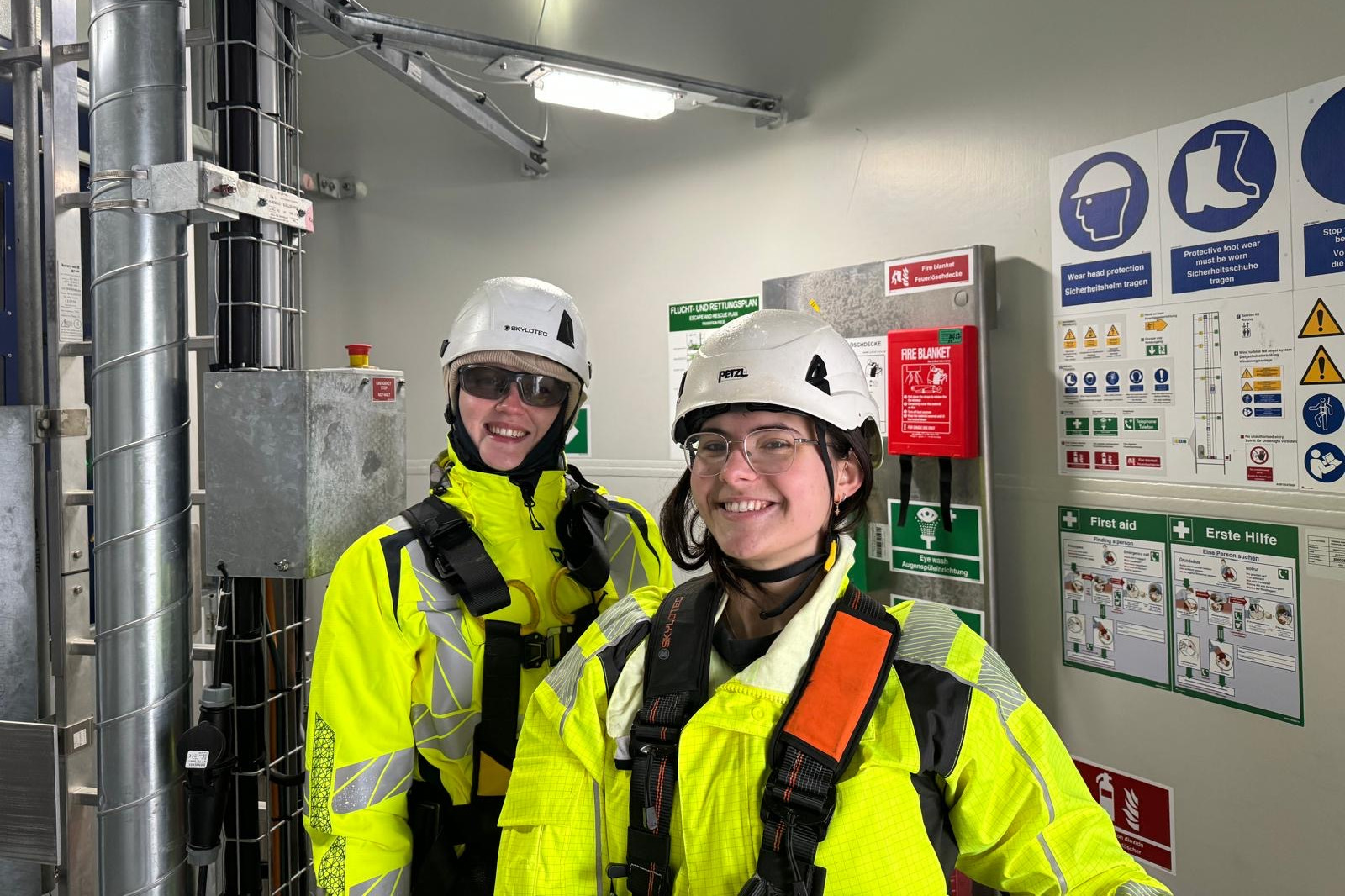 Two safety workers in bright yellow jackets and helmets stand in a facility, surrounded by safety signs and equipment.