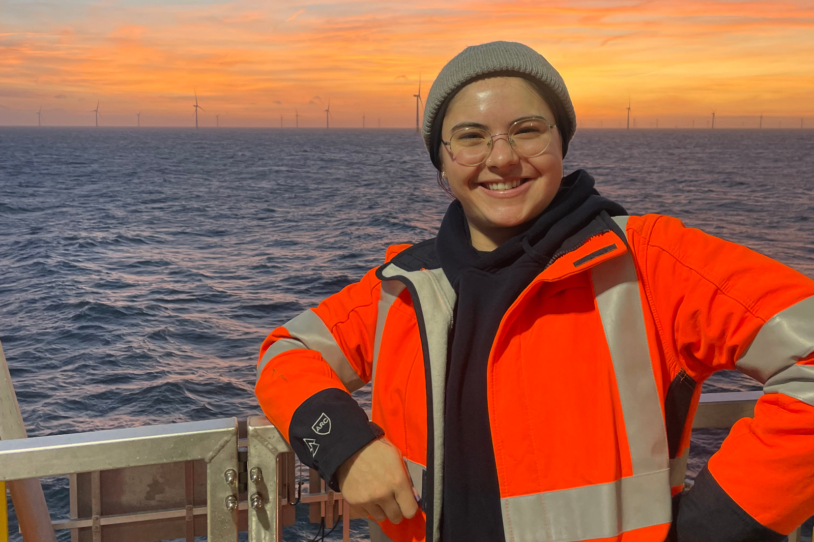 A person in an orange safety jacket and grey beanie stands on a ship's deck, with wind turbines visible in the background at sunset.