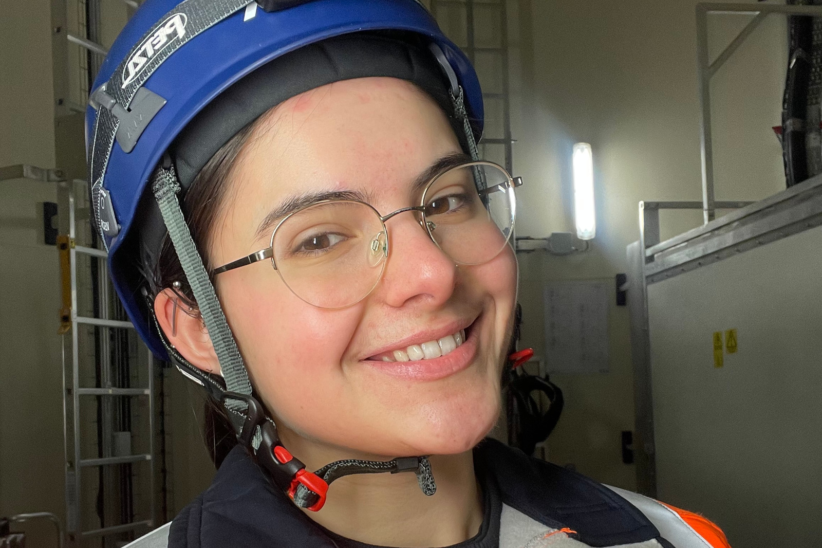 A person wearing a blue helmet and an orange safety vest standing in a well-lit industrial environment.