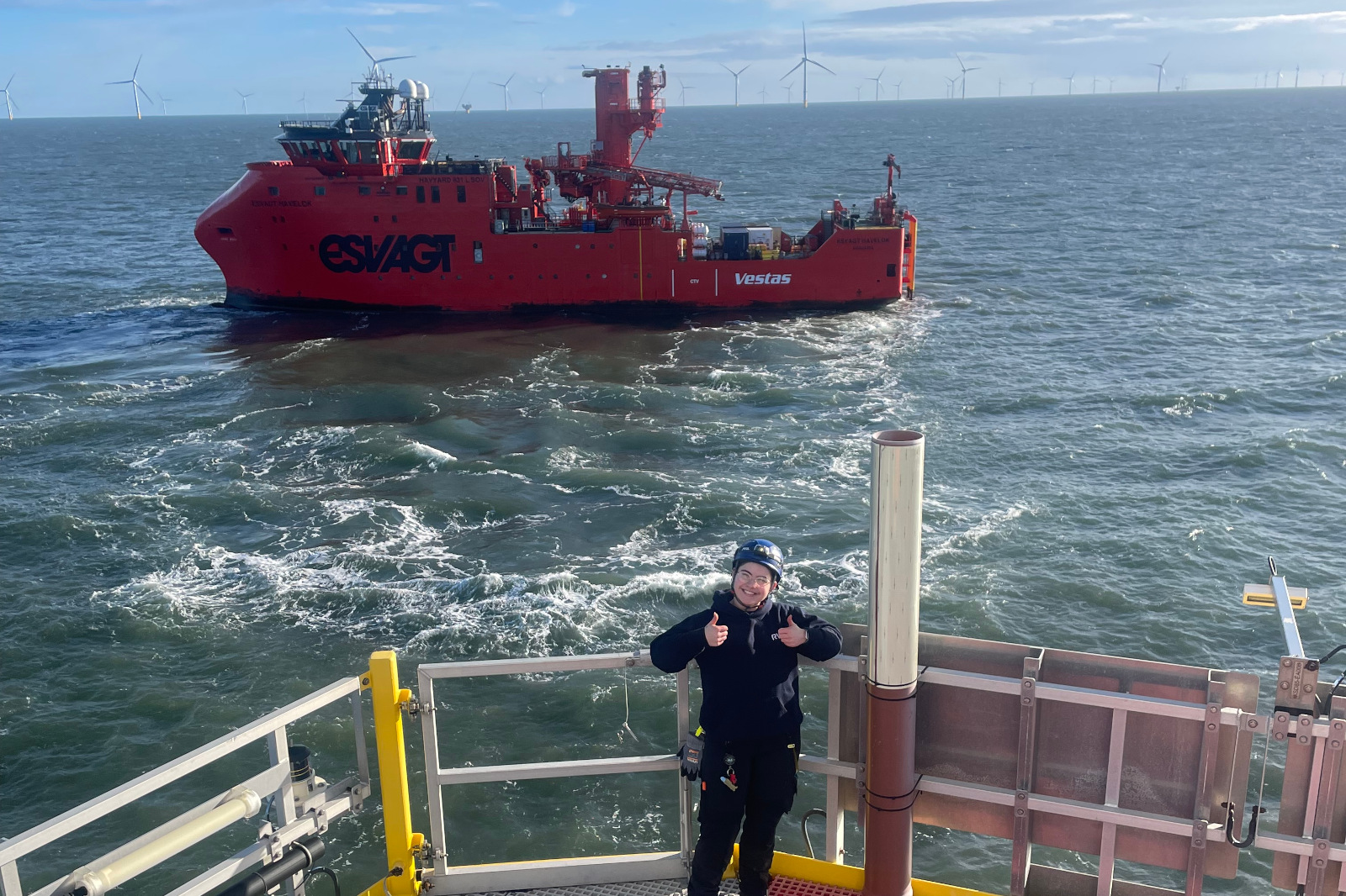 A red vessel sails through choppy waters, with wind turbines in the distance and a crew member giving a thumbs up.