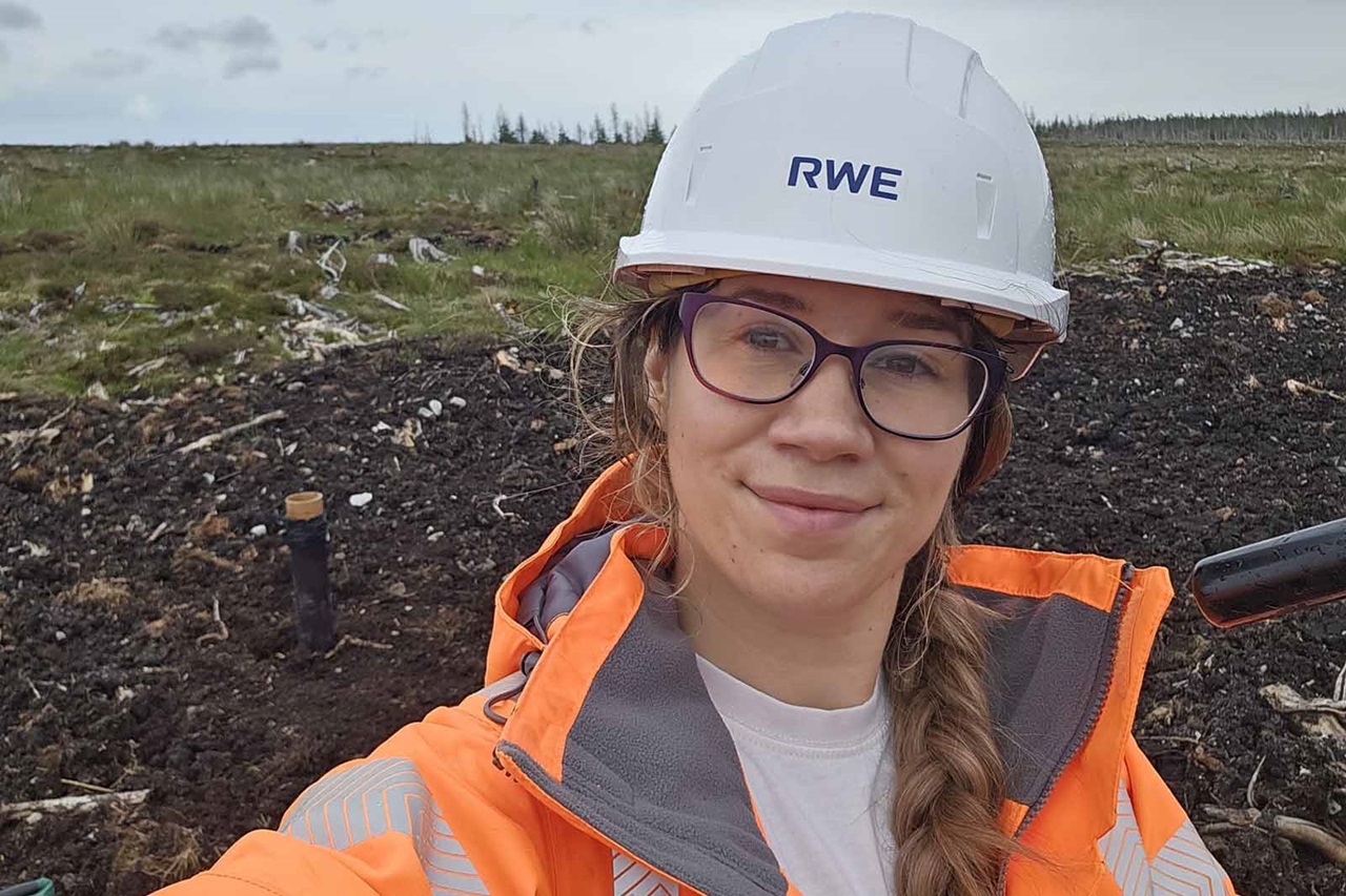 A person in a white hard hat labeled "RWE" and an orange high-visibility jacket smiles slightly in a grassy peatland under a gray overcast sky. Portrait of Heather Johnstone.