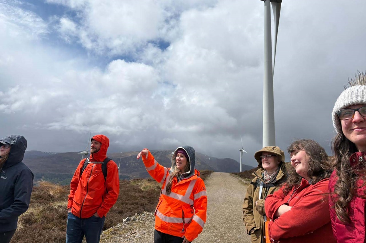 A group of people in orange, red and tan jackets stand on a gravel path beside a tall white wind turbine under cloudy gray skies.