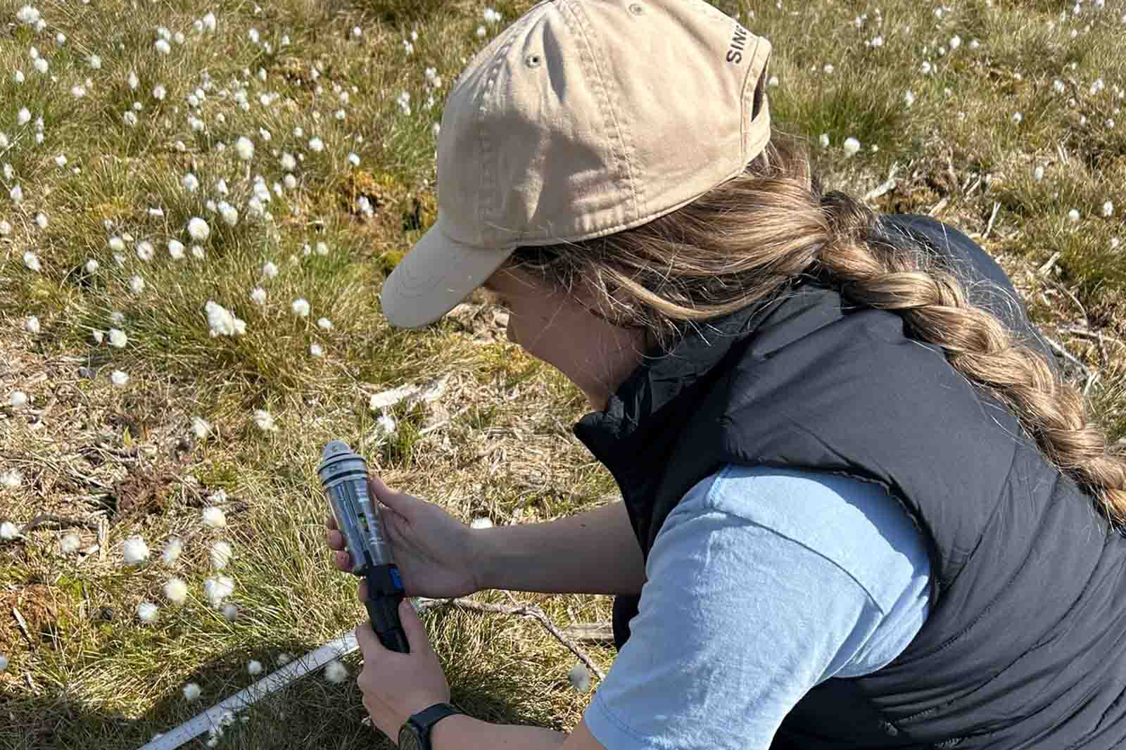A person wearing a beige baseball cap, light blue shirt and black vest kneels in a grassy field dotted with white cottonlike seed heads, holding a small metal instrument and a coiled white rope.