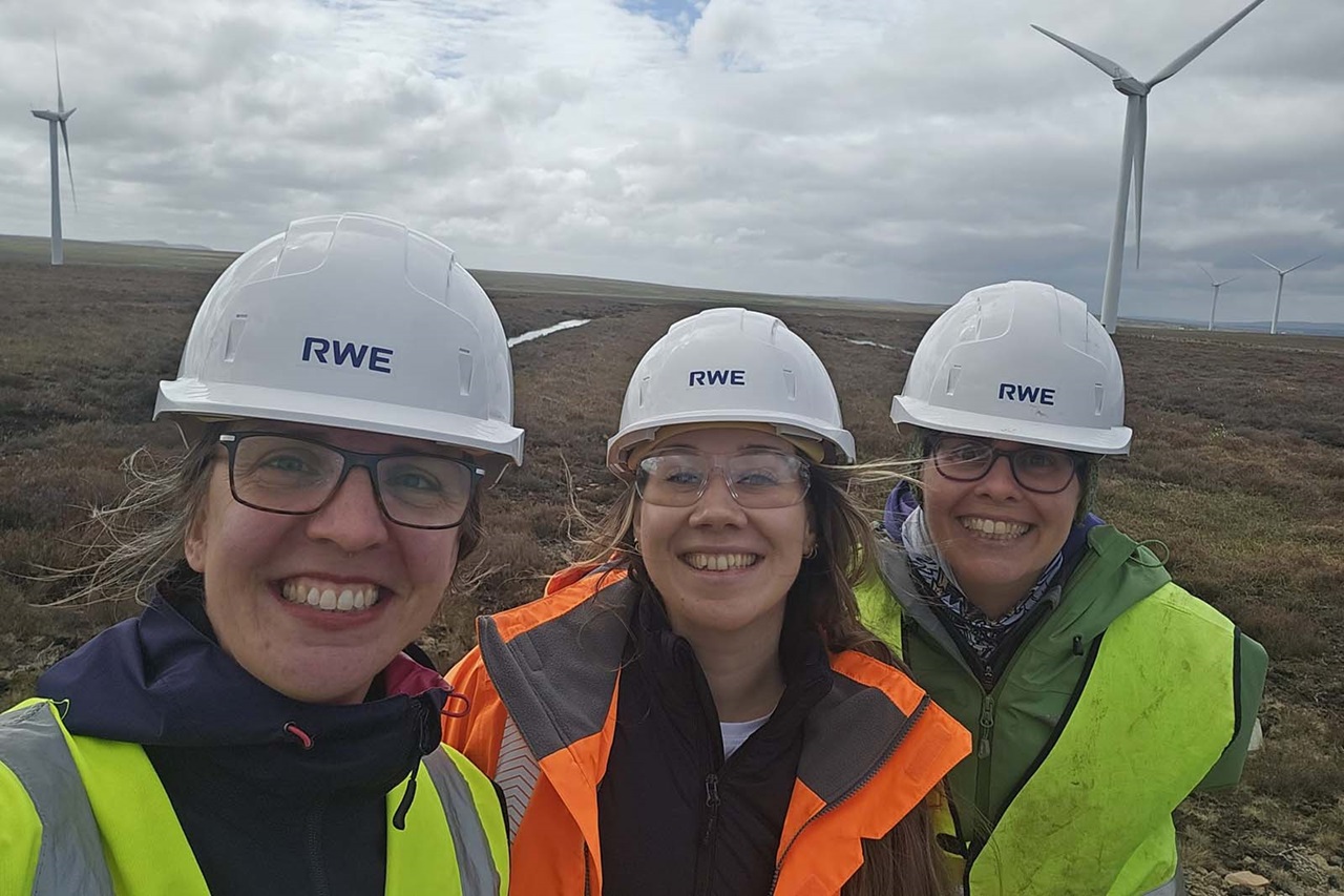 Three smiling workers wearing white hard hats labeled "RWE", safety glasses and bright yellow or orange vests stand on heather moorland with wind turbines.