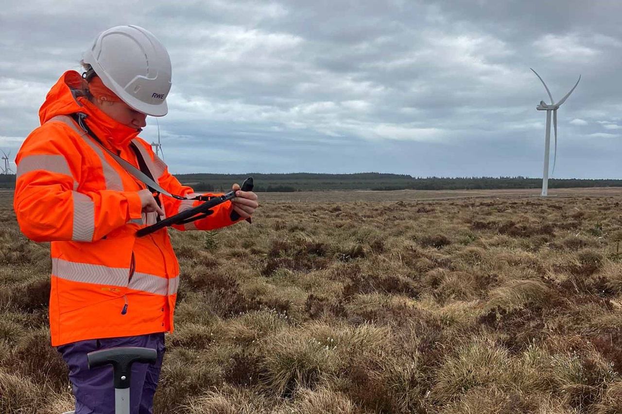 Person in a bright orange jacket and white hard hat stands in brown moorland holding a tablet, a white wind turbine visible under an overcast sky.