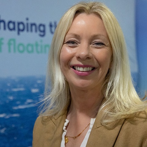 A woman with long blonde hair wearing a beige blazer and necklace, standing in front of a backdrop featuring blue ocean waves.