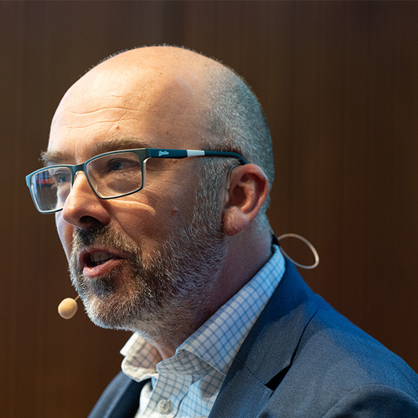 A man in a blue suit jacket and checkered shirt wears glasses and a behind-the-ear microphone against dark wood-paneled wall. Picture of Jon Page.