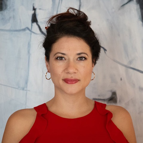 A woman in a red top with brown hair in updo style stands again neutral background. Portrait of Alice Barrs.