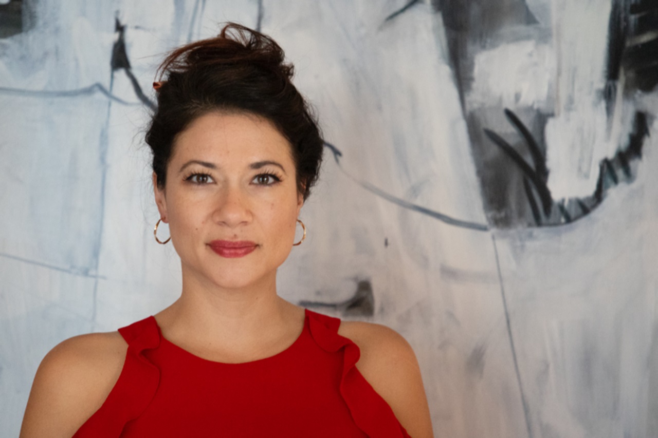 A person wears red sleeveless top with ruffle shoulders and gold hoop earrings, hair in bun, face covered by neutral square against abstract mural.