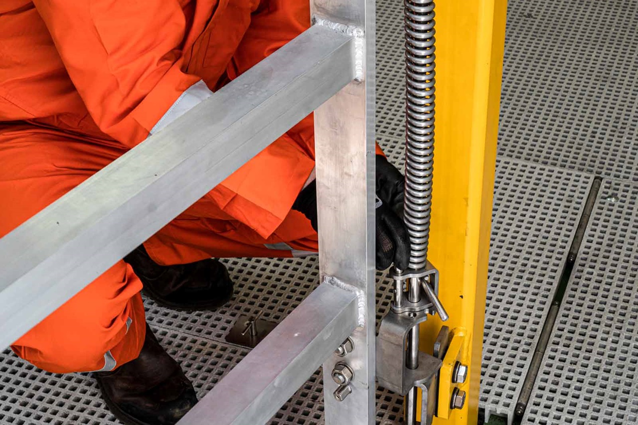 A worker in an orange suit adjusts metal scaffolding components while kneeling on a grated platform.