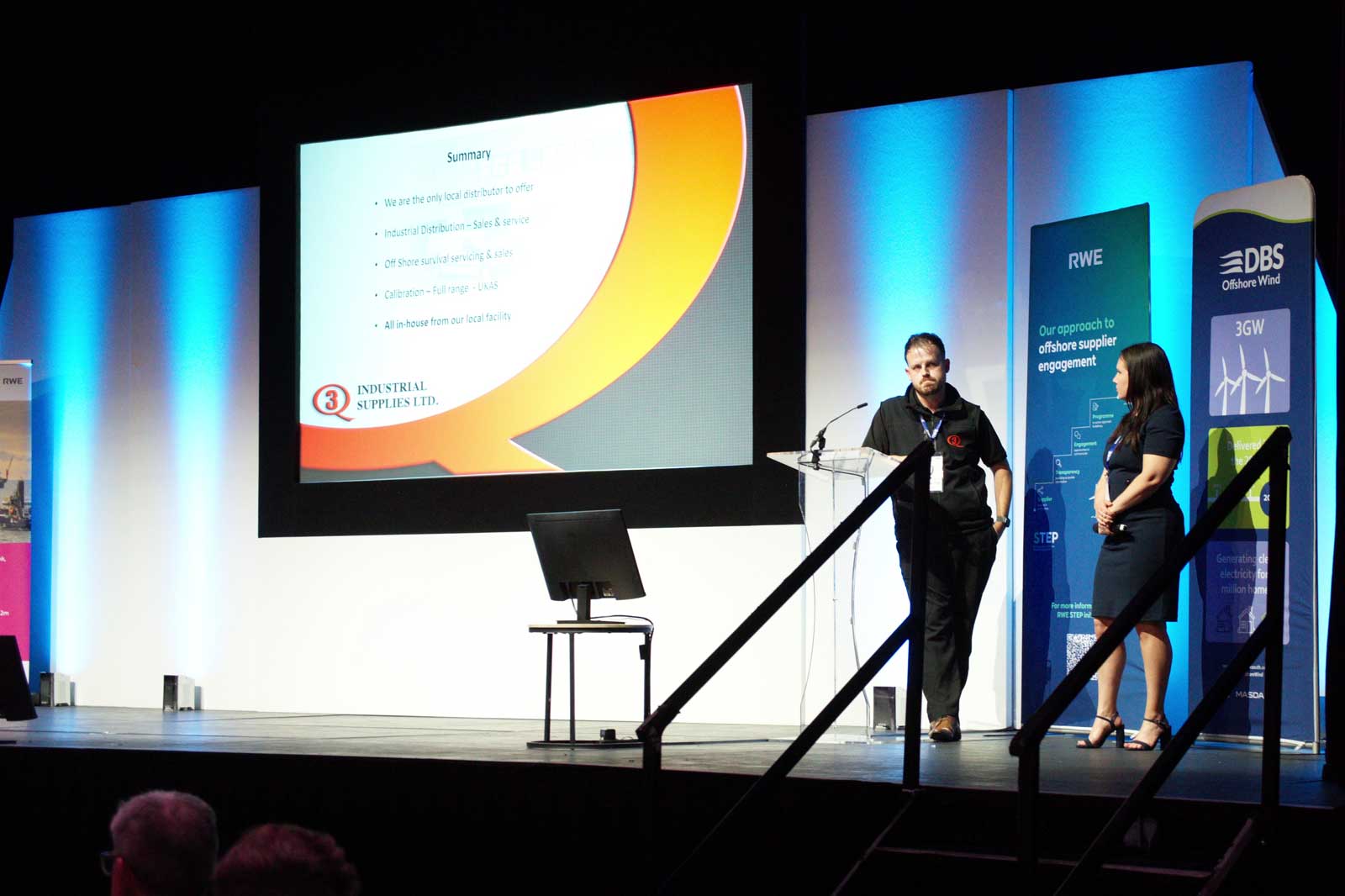 A presentation scene featuring two speakers beside a screen displaying key points about industrial supplies and offshore servicing.