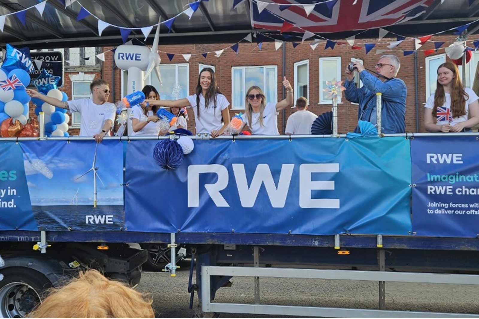 A festive event with people on a decorated float, holding flags, balloons, and signs for RWE, celebrating community spirit.