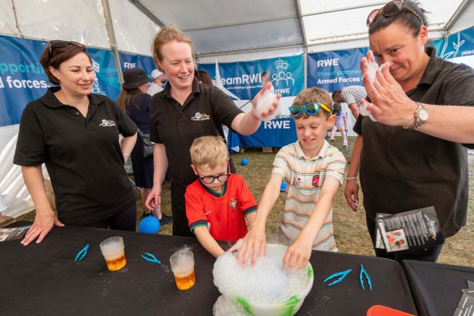 A child in a red shirt plays with bubbles at a festival stall, while adults in black shirts assist.