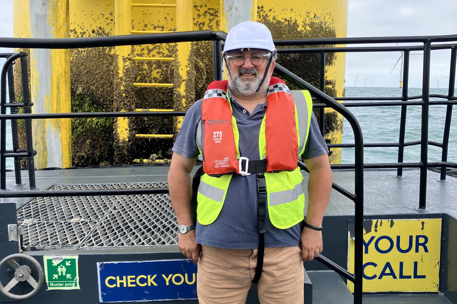 Man wearing white hard hat, orange lifejacket and neon-yellow vest stands on a metal deck by a yellow structure; signs read "CHECK YOUR" and "YOUR CALL".