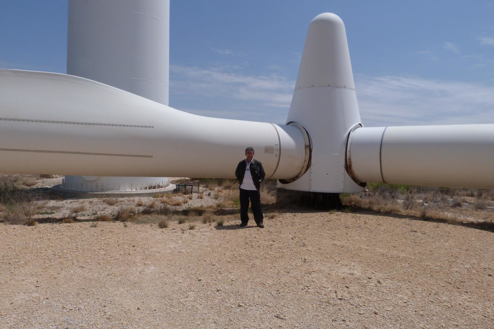 A man in dark clothing stands beside a massive white metal wind turbine hub and blades on pale gravel under a clear blue sky.
