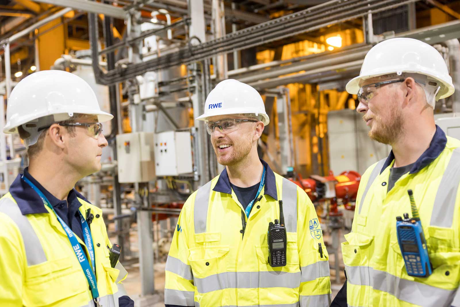 Three workers in safety gear converse in an industrial setting, with machinery and control panels in the background.
