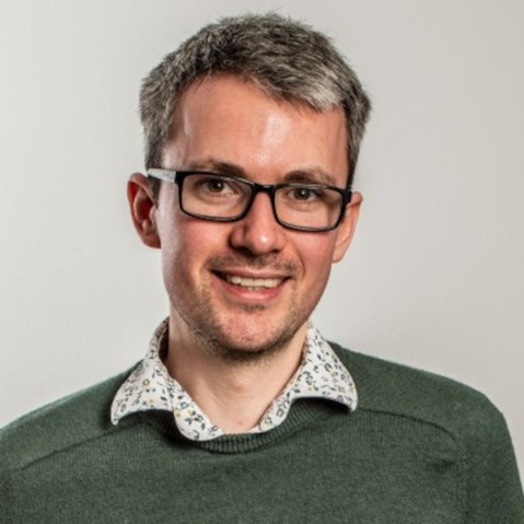 A person from the shoulders up with short salt-and-pepper hair wearing a green sweater and a white collared floral shirt against a light background.