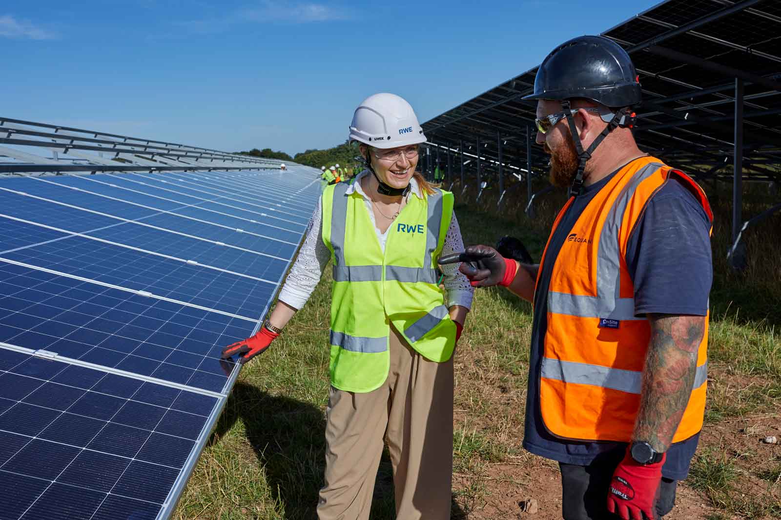 Two workers discuss solar panels in a field, wearing safety gear. The sun shines brightly, and many panels are visible.