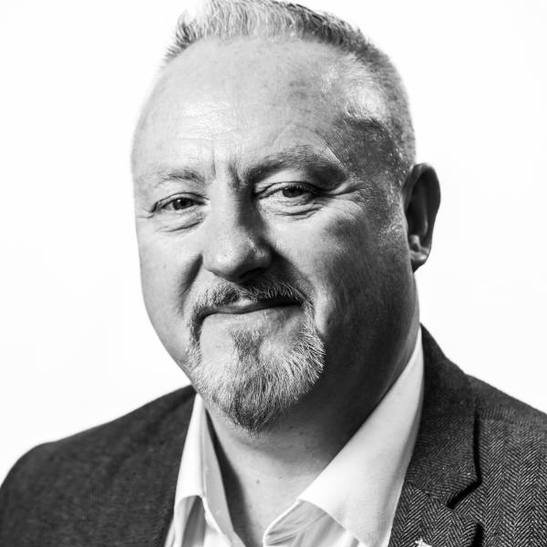 A man in a grey blazer with a white shirt, set against a plain white background.