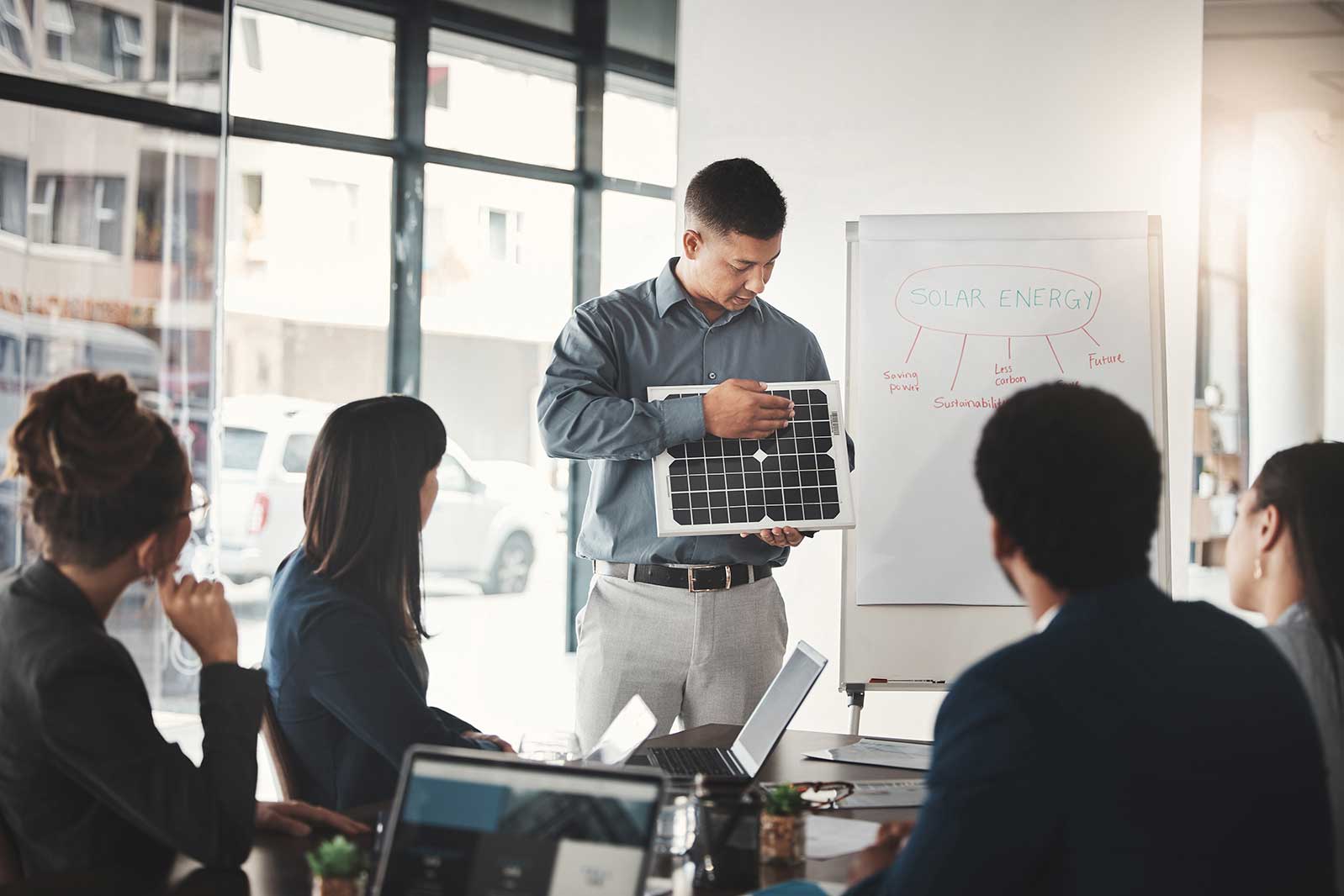 A business meeting where a presenter shows a solar panel while discussing solar energy concepts with attentive colleagues.