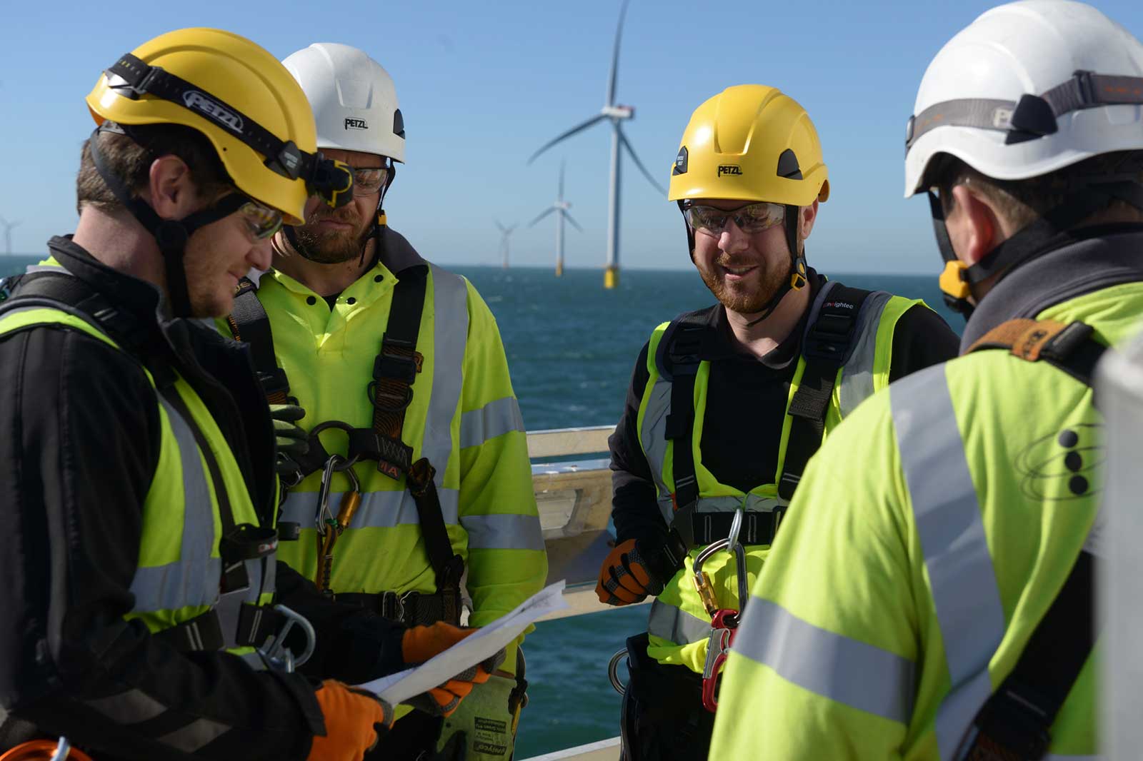 Four workers in safety gear discuss plans on a platform, with wind turbines visible in the background.