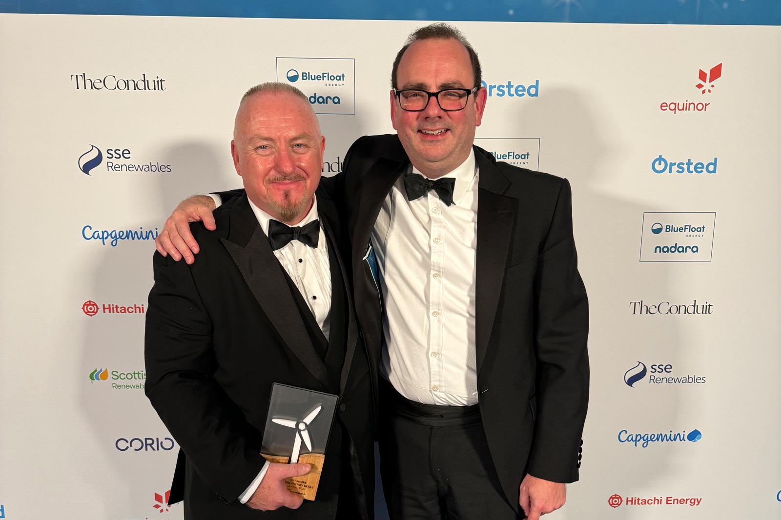 Two men in black tuxedos pose together, one holding a trophy with a wind turbine design, at an awards event backdrop.