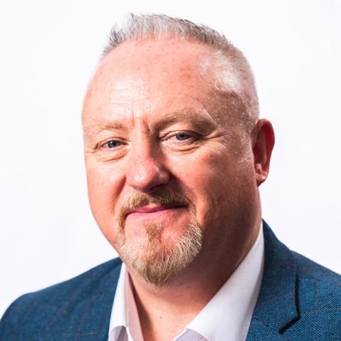 A person wearing a blue blazer over a white shirt, with short hair and a beard, set against a plain white background.