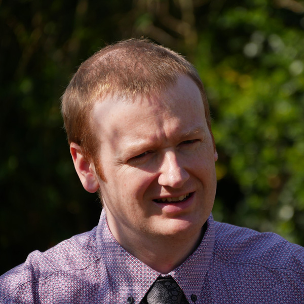 A man wearing a patterned shirt and tie is seated outdoors with greenery in the background. Portrait of Kester Gunn.