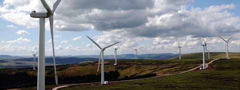 A panoramic view of multiple wind turbines on a rolling landscape under a partly cloudy sky.