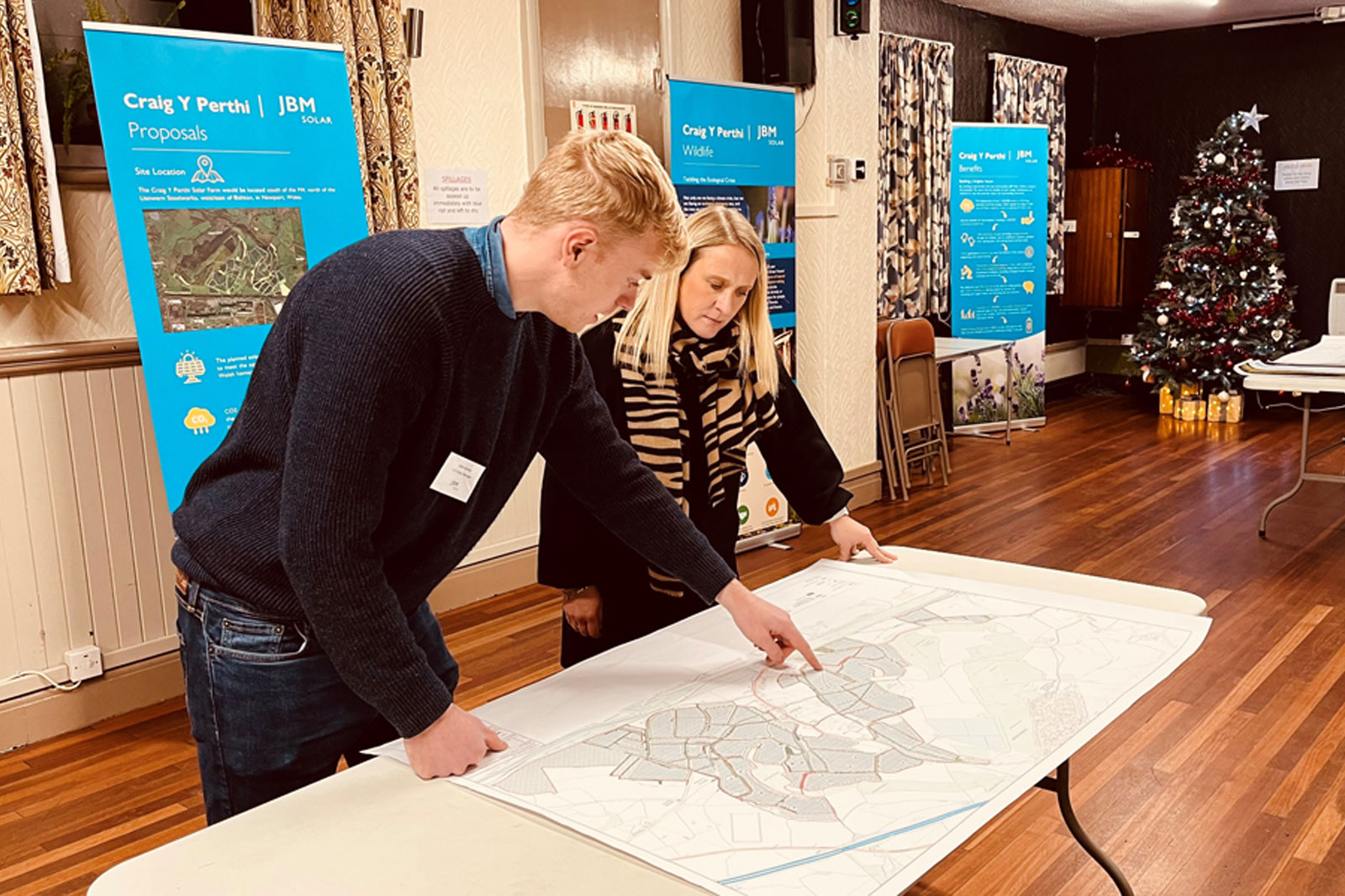 Two individuals examining a map on a table at an event with information boards and a Christmas tree in the background.