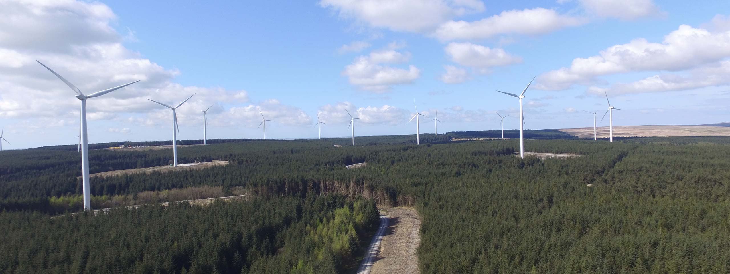 Aerial view of wind turbines in a green forest under a blue sky with clouds.