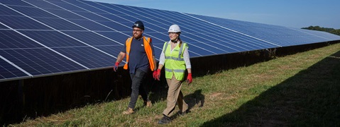 A man in an orange vest and a woman in a reflective jacket walk among solar panels under a clear blue sky.