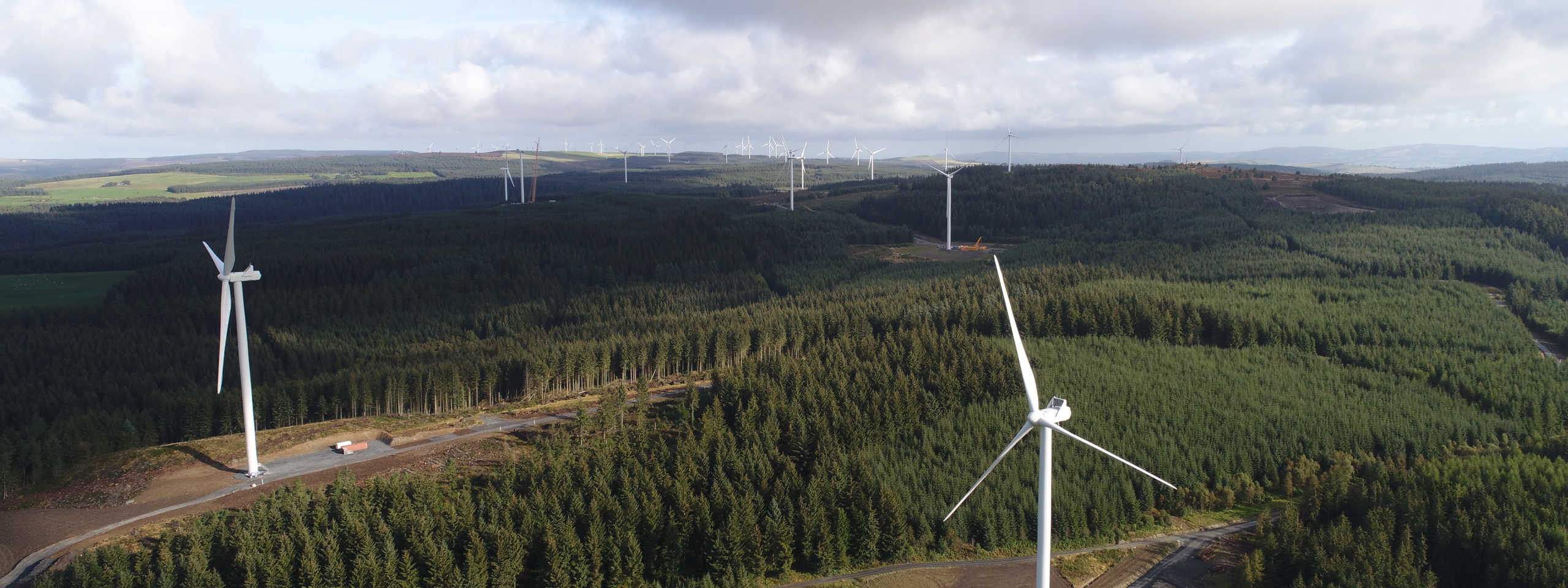 Aerial view of wind turbines in a lush forested area, with hills in the background under a partly cloudy sky.