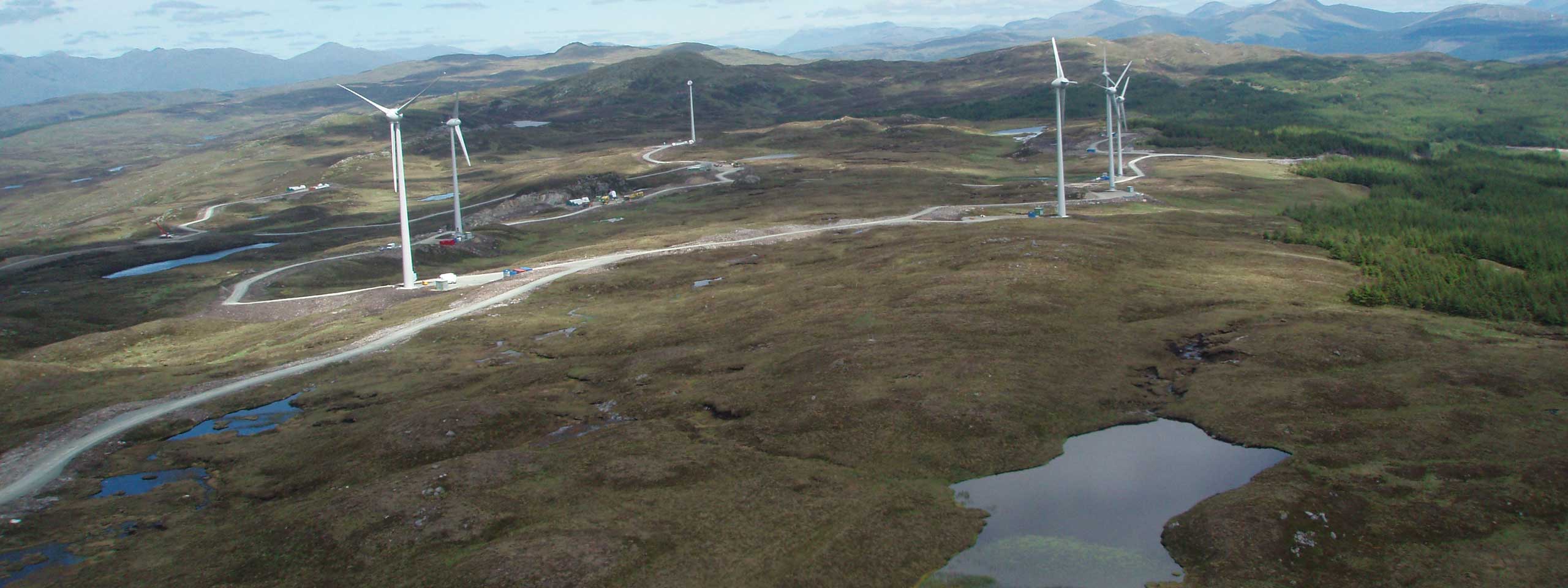 Aerial view of a landscape featuring wind turbines, winding roads, and patches of water amid hills and greenery.