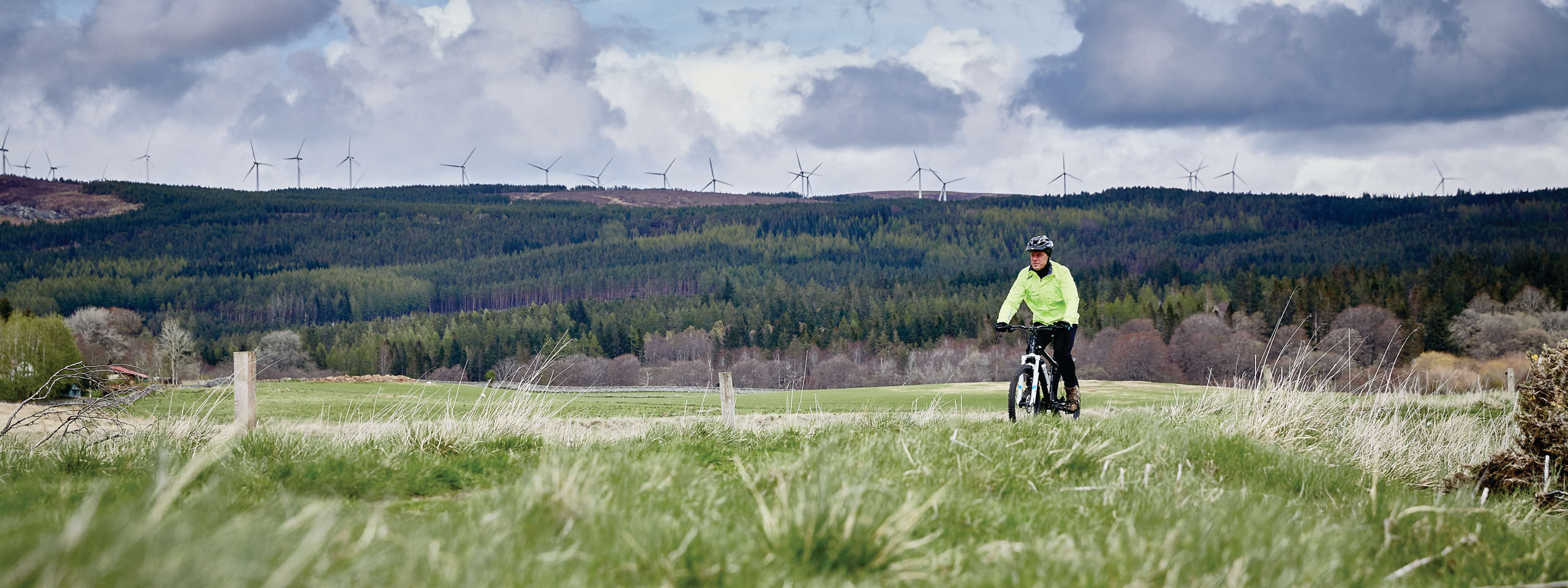 A cyclist in a bright yellow jacket rides through a grassy field with wind turbines in the background and rolling hills.