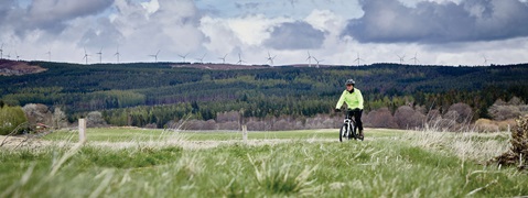 A cyclist in a bright yellow jacket rides through a grassy field with wind turbines in the background and rolling hills.
