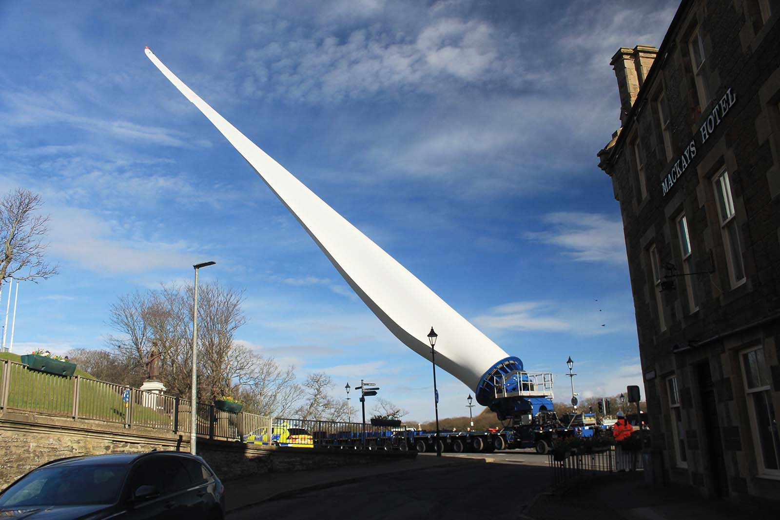 A massive white wind turbine blade dominates a street, mounted on a heavy trailer beside stone buildings under a bright blue sky.